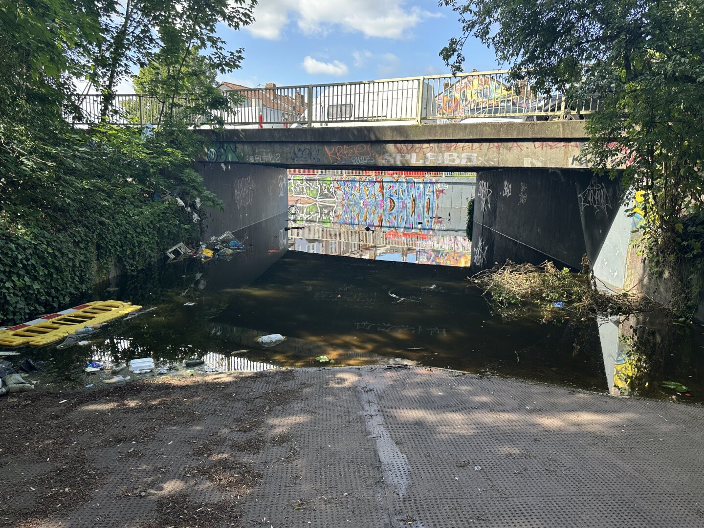 A pedestrian underpass under a road. It is overgrown on either side, and there is a street art mural visible in the distance on a concrete wall. The underpass is completely full of water with some random items in it. It's depth is not visible but in the centre it looks at at least 25 cm deep. The water is still under the mural is reflected.