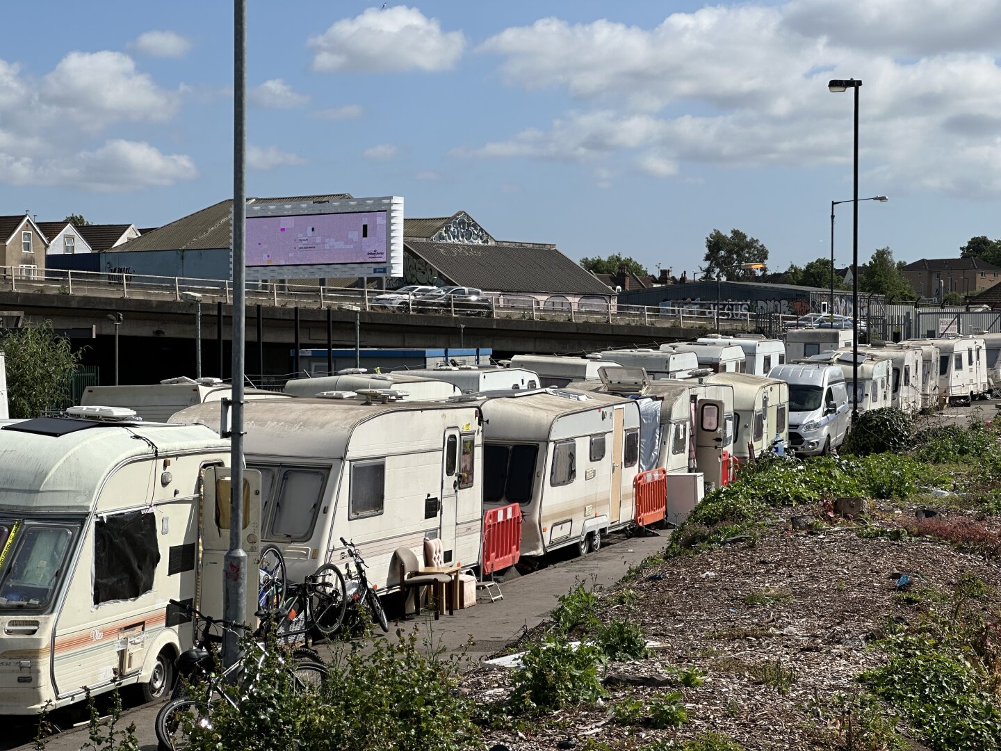 Who wrote Line on either side by dilapidated caravans. These have not moved for sometime; there are chairs outside one of them and bicycles leaning against others. Behind and above the caravans is a motorway flyover with some cars. Behind that is a billboard advertising products for the drivers and passengers.
