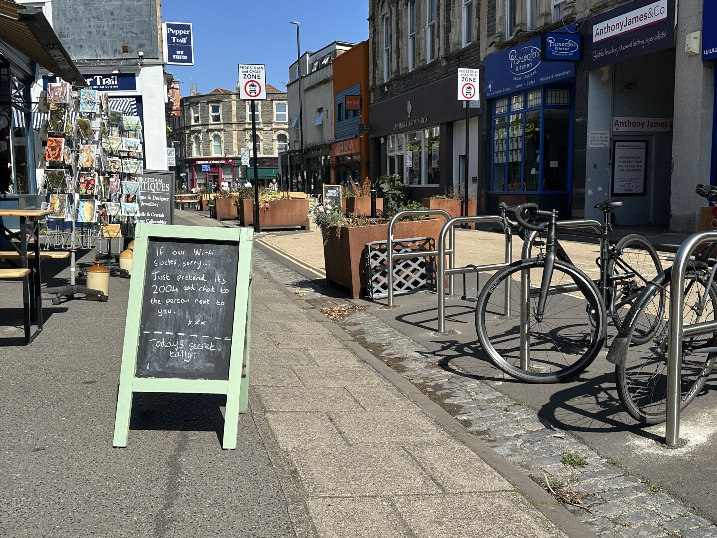 A street scene featuring outdoor seating, a sandwich board sign with a humorous message about Wi-Fi, and a display of magazines. Bicycle racks with bikes are visible in the foreground, while shops line the street in the background. The area is sunny and pedestrian