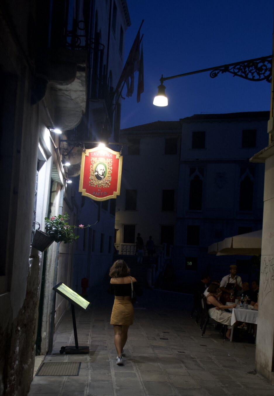 A night scene with a dark blue sky. On a street in Venice, a woman walks under a street light. To her left is an open restaurant whose light also illuminates her side. In front of the restaurant is a red and gold sign.