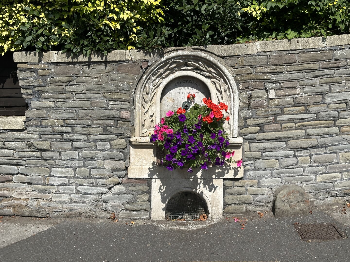 An ornate stone fountain embedded in a wall is adorned with vibrant flowers, including red and purple petunias. The surrounding area features lush green foliage, providing a picturesque setting.