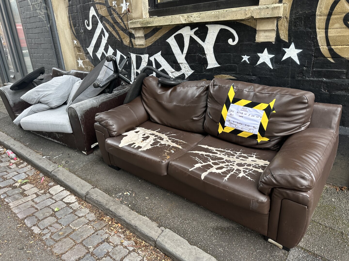 Two discarded sofas are seen beside a wall with a mural. One is brown leather with visible wear and a sign labeled "FREE" attached, while the other is a grey fabric couch with cushions piled on it. The area is in an urban setting,