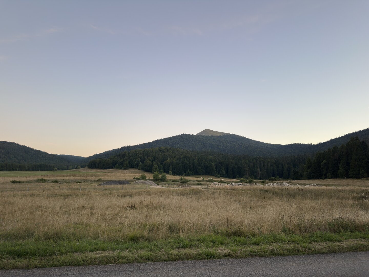 A sky at the sunset with wooded hills beneath. In the foreground is a field of grass.
