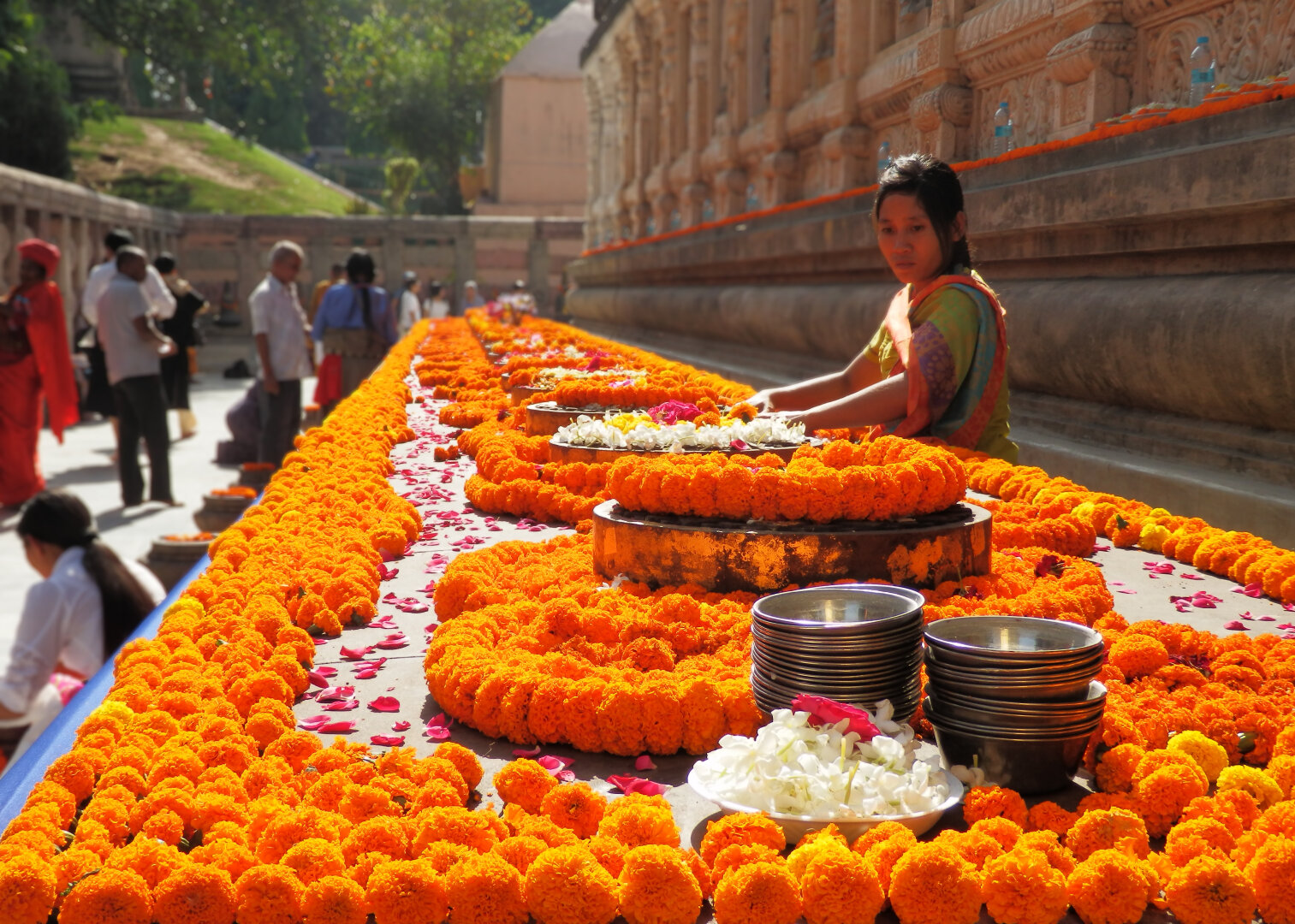 The image showcases a vibrant and colorful scene at Mahabodhi temple in Bodh Gaya, India. The primary focus is on a long, meticulously arranged display of bright orange marigold garlands. A woman, dressed in a colorful sari, is the central human figure. She is positioned behind the marigold display, and is arranging the flowers. On the left side of the flower is an out-of-focus crowd of people.