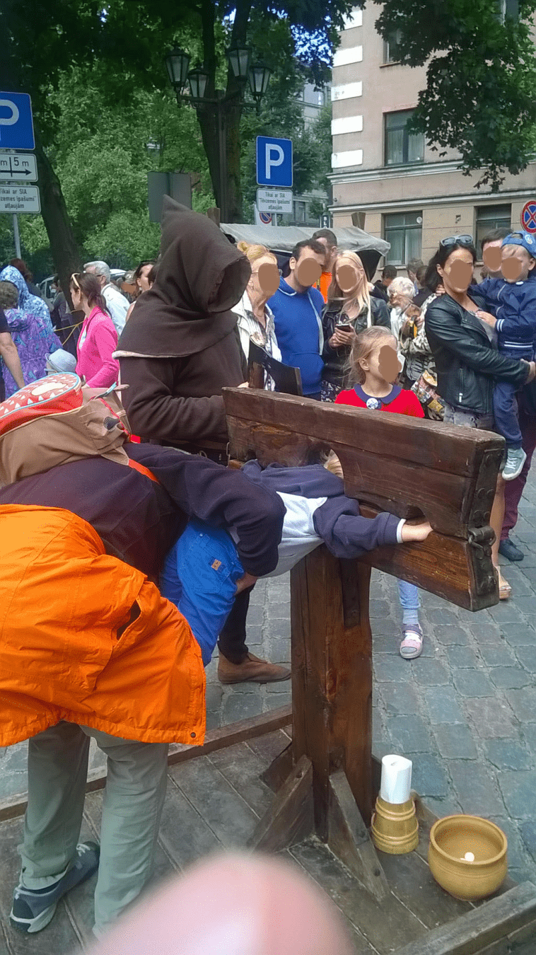 A very young boy is in the stocks, being lifted to fit into the stocks