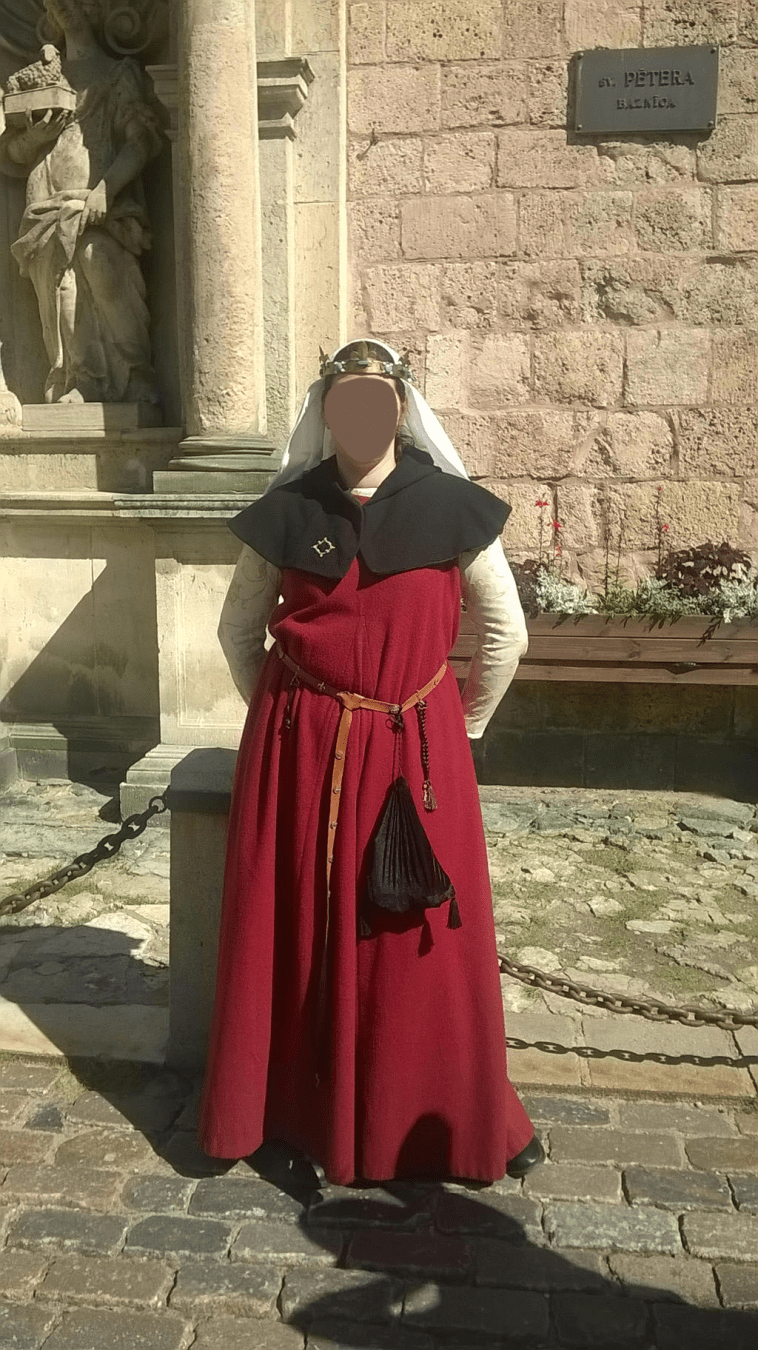 A young woman, dressed in traditional medieval clothing, standing in front of the church wall, hiding behind her back her lunch from a corner store