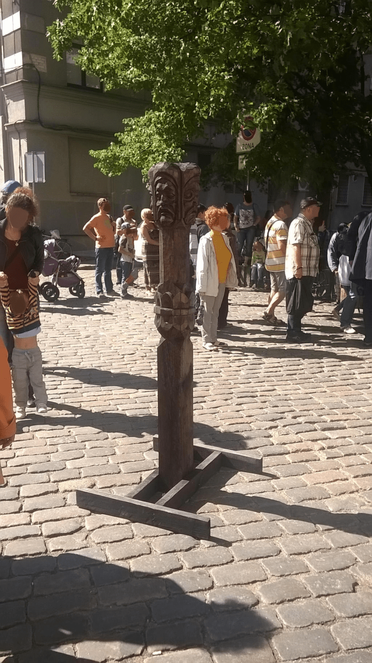 Photo of a wooden pillar, placed in the middle of the church square, with people milling about the square