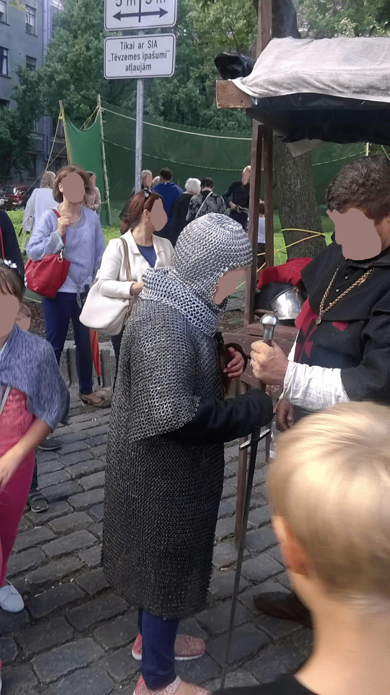 A youth dressed in mesh armour and being given a sword by a fair worker