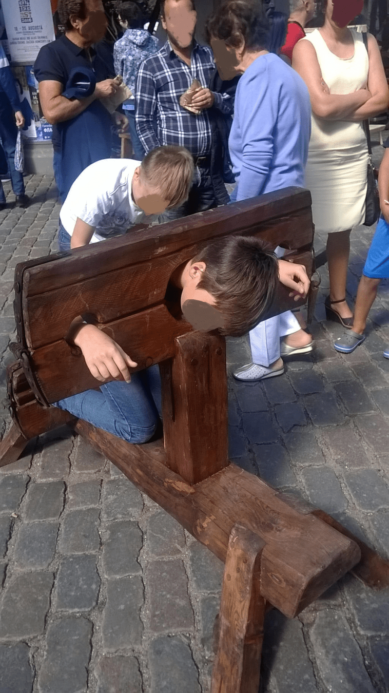 A boy in the stocks with another boy helping him into it, with people around the area