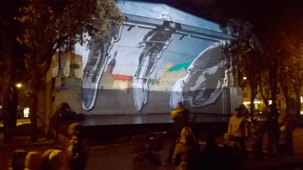 Cutouts of cyclists racing across the background that is a painting of old Riga