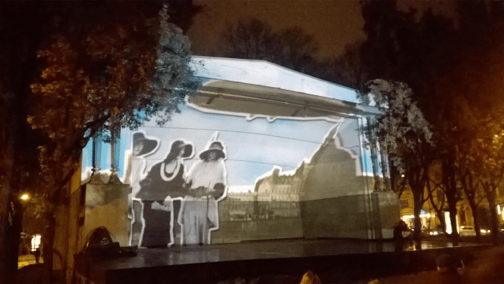 Black and white cutouts of ladies, standing in front of a yellow-ish cutout of the city of Riga, with a yellow-ish cutout of a dirigible flying over head in the blue skies