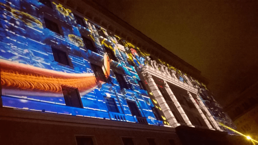 Front facade of a hotel, lit up by a light display, with blue flames, yellow flowers and a long shape of some kind appearing on it