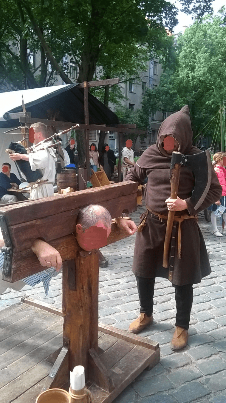 An older man is in the stocks, but is a bit bigger than the stocks were meant for, so they don't lock fully