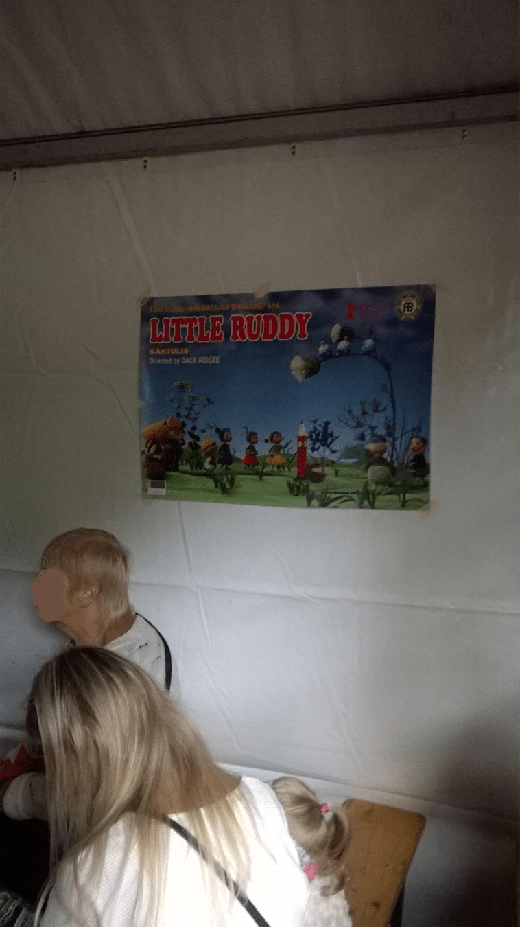 Photo from inside of a different tent, with people watching a movie, focus on the poster for the movie "Little Ruddy"