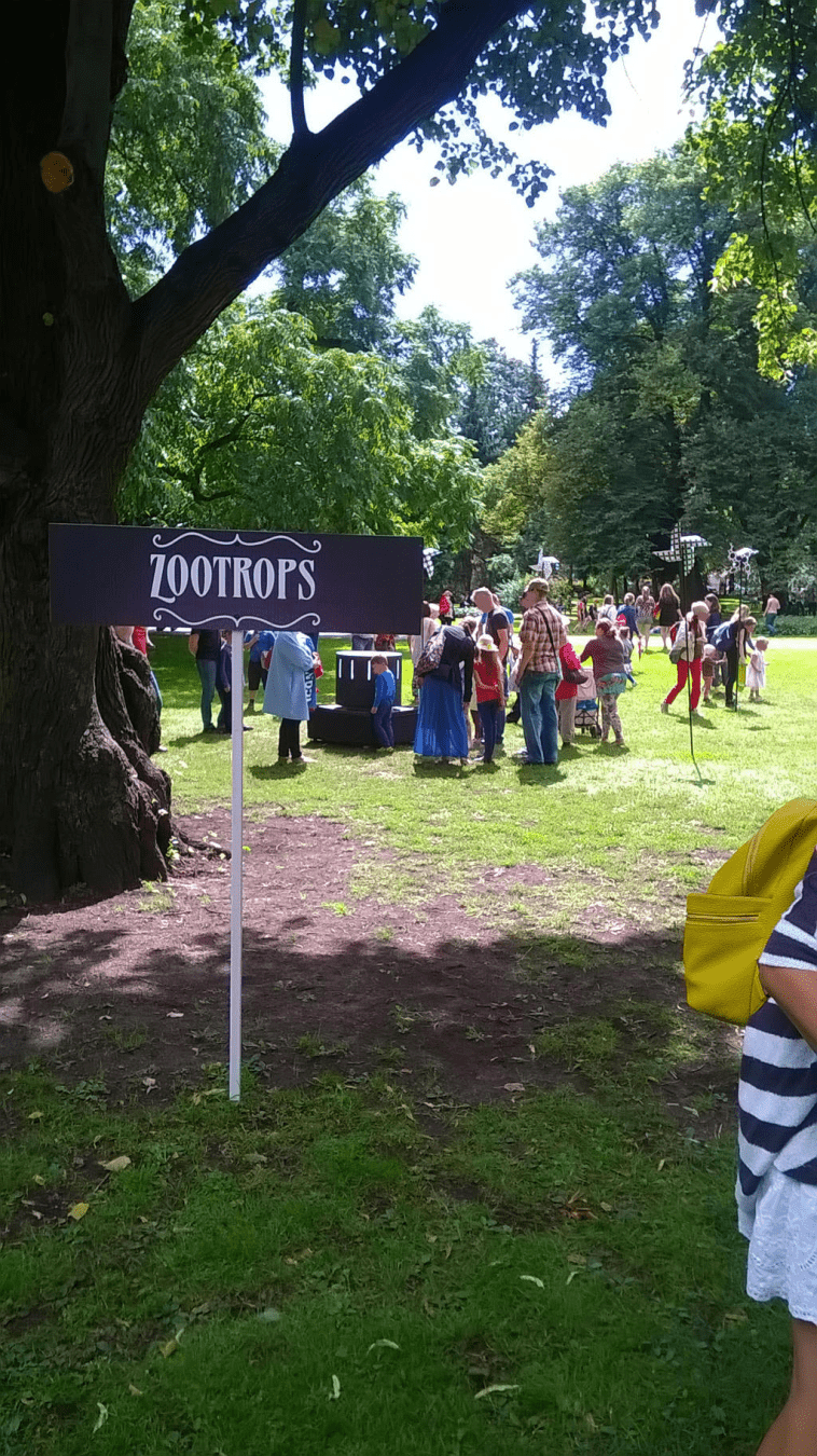 Photo of the sign for the Zootrops in the summer fair, with people around the titular zootrop