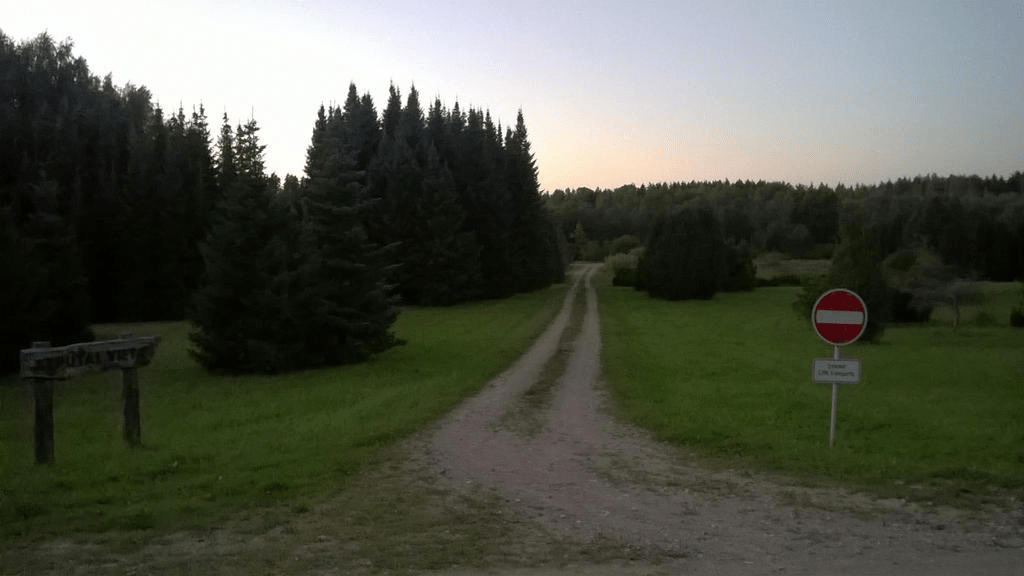 Dirt road through the lush grass with patches of trees and bushes scattered around, a wooden sign on the left and a stop sign on the right