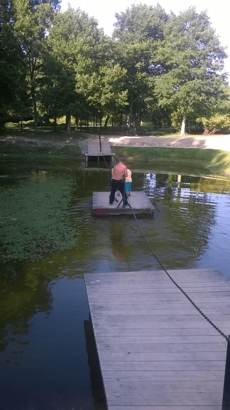 Photo of a man and young boys pulling themselves across on a wooden plank with chains from one dock to the other across the pond
