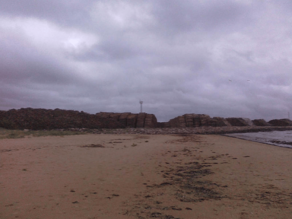 View of the beach, with a wood mill type area in the distance, with the sea to the right, the sky covered by thick clouds
