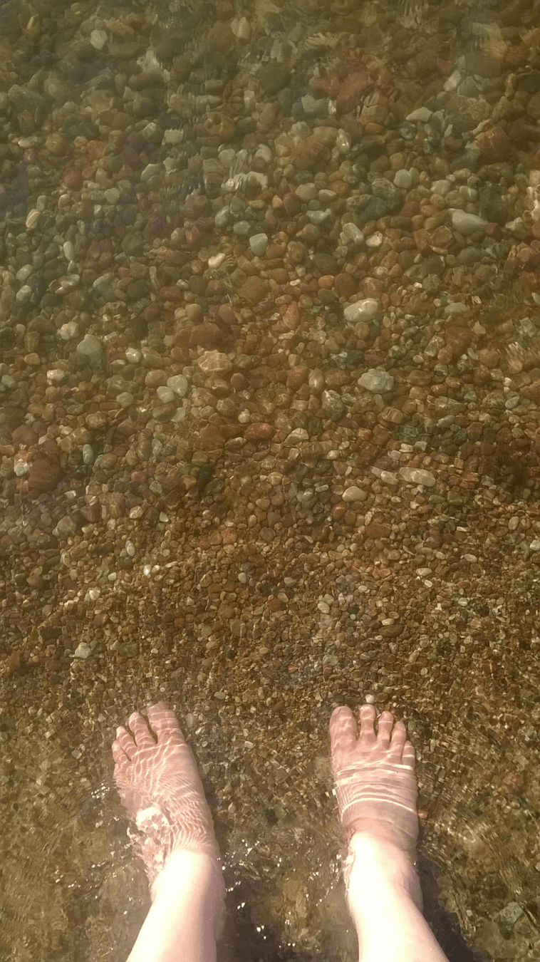 Pebbled ground in the shallows of the sea at a beach, with human feet in the water at the bottom of the picture