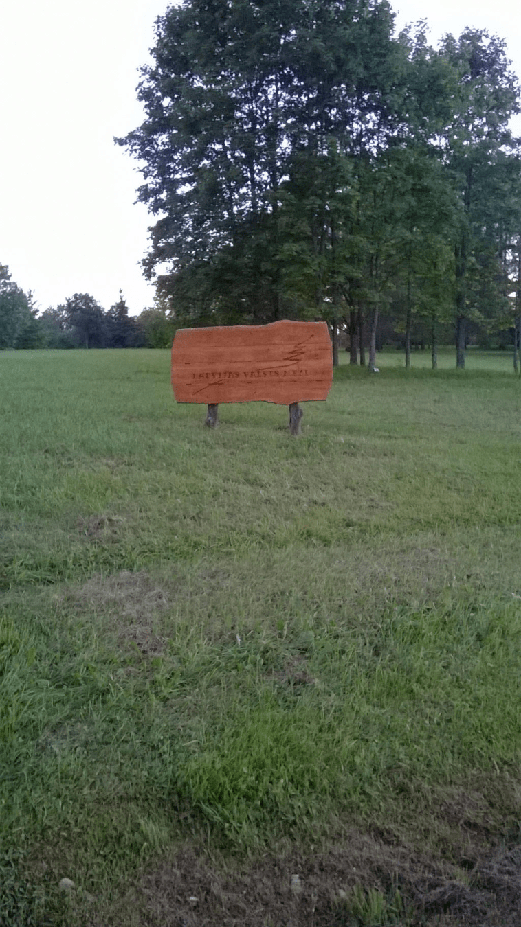 A wooden sign of the Latvian Forests on a small hill with cut grass around and trees in the distance
