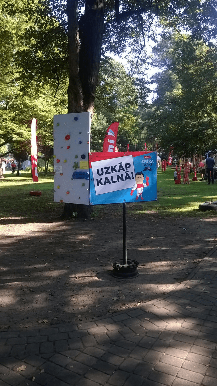 A sign that challenges you to climb a climbing wall, one in the background attached to a tree...