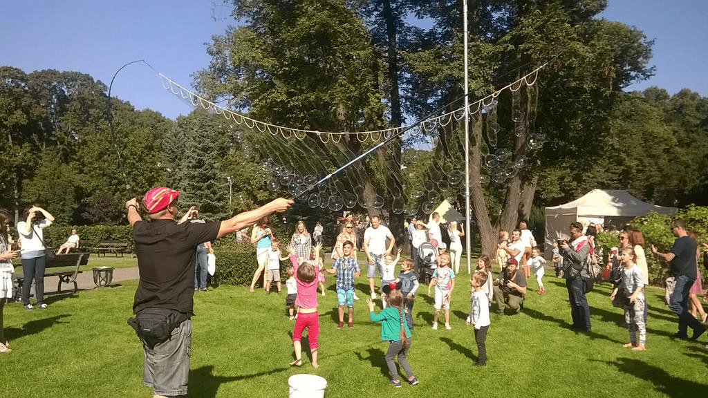 Man making more bubbles for small kids with a soapy rope between two fishing poles during a bright summer day during a fair