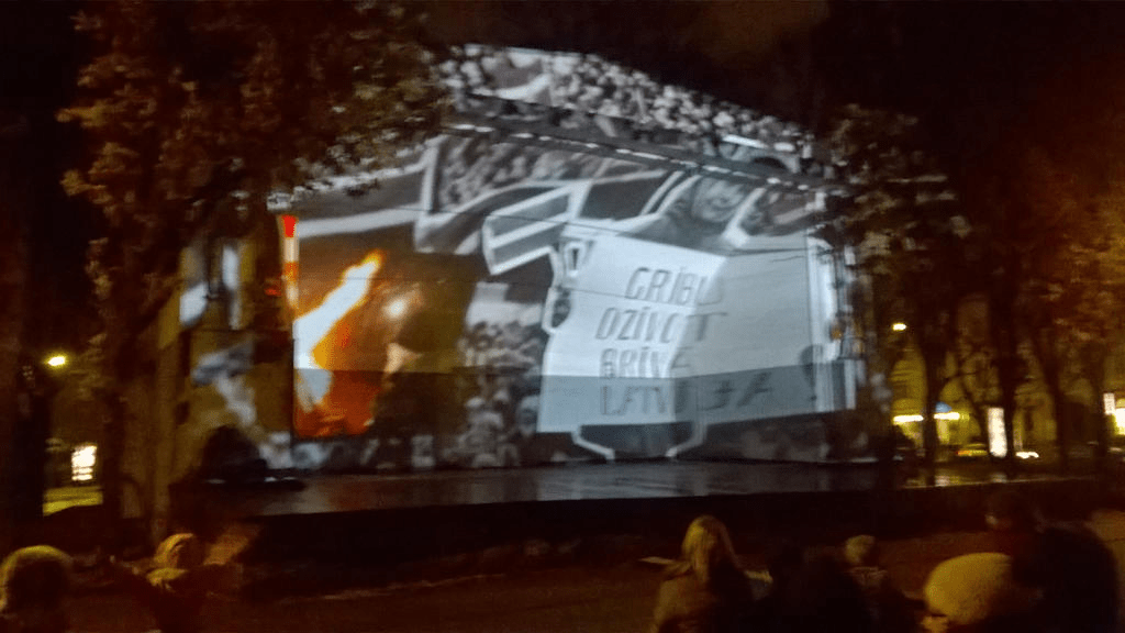 The march, with a clip of fire in the corner and a cutout of someone holding a sign saying in Latvian that they want to live in a free Latvia