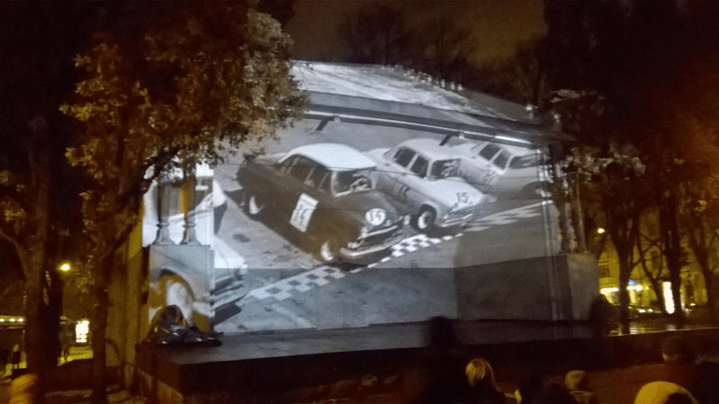 Still from a black and white clip, showing old cars readying for a race at the start line