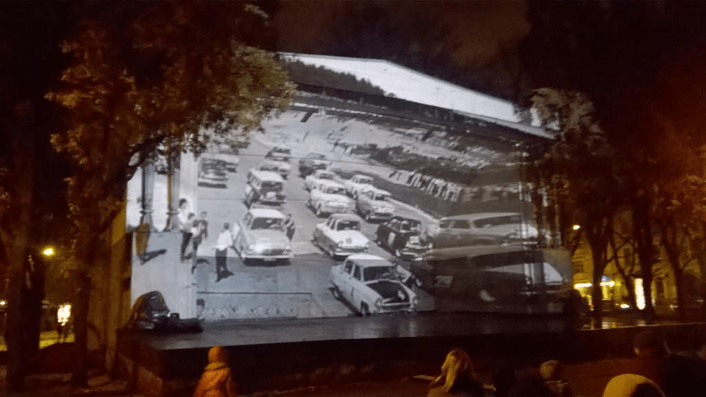 Still from a black and white clip, with old cars and people gathering