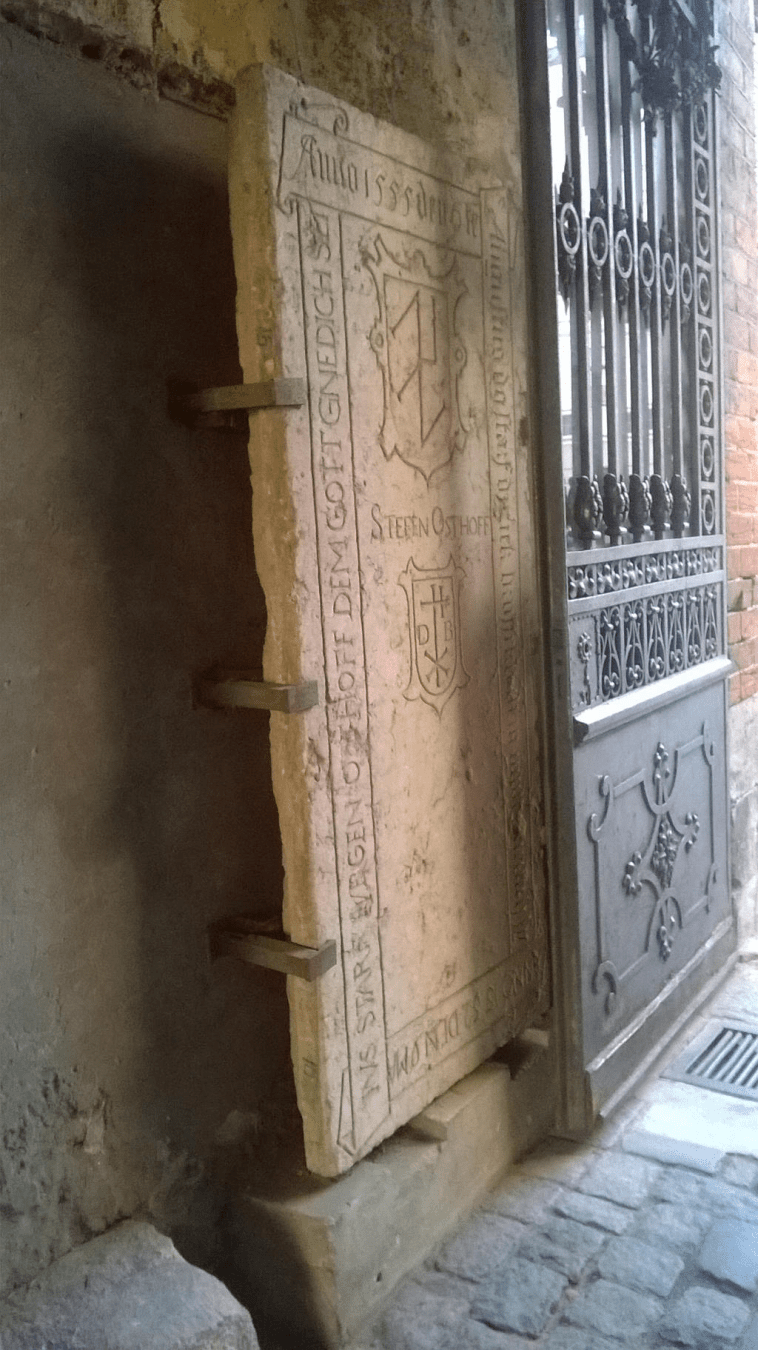 Photo of another stone door, with Latin writing on the sides and coats of arms (upper larger than the lower one), held to the wall by metal clamps.