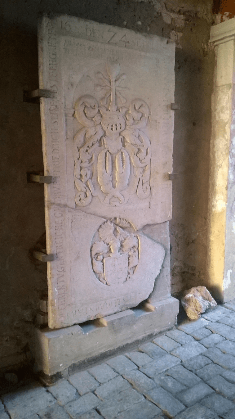 Photo of a broken stone door, with decorative design (lower part of a very fancy coat of arms and upper part looks like a flowery pot coat of arms), with Latin writing on the sides of the door, held to the wall by metal clamps.