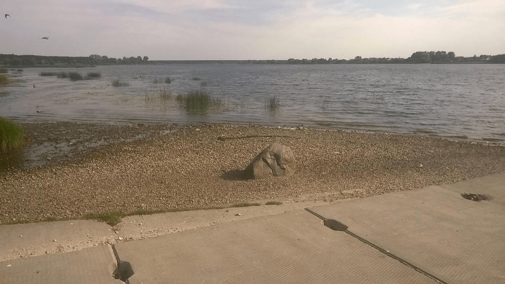 Photo taken from an old dock on a river. The visible part of the old dock is made of rock, the ground going into the water is covered in small stones, with a larger stone sitting in the middle of it, with water receding from its normal location. Water is calm, with reeds growing in it closer to dry land.