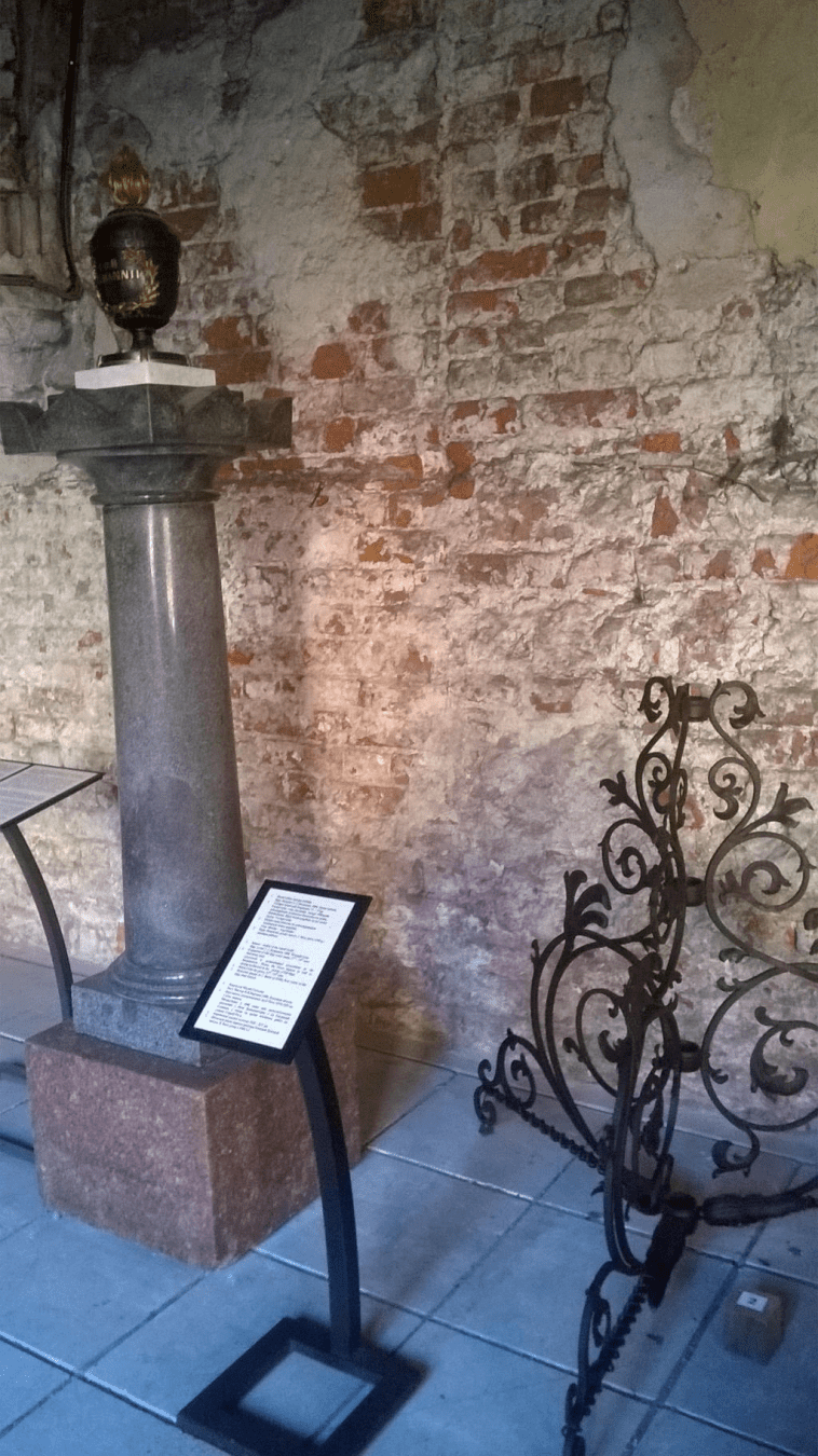 Metallic decoration on the ground, made to look like vines going up (meant to hold something on fire?) and a stylish stone pillar on a stone block and a metallic goblet of sorts atop of the pillar, with dark metal and gold text
