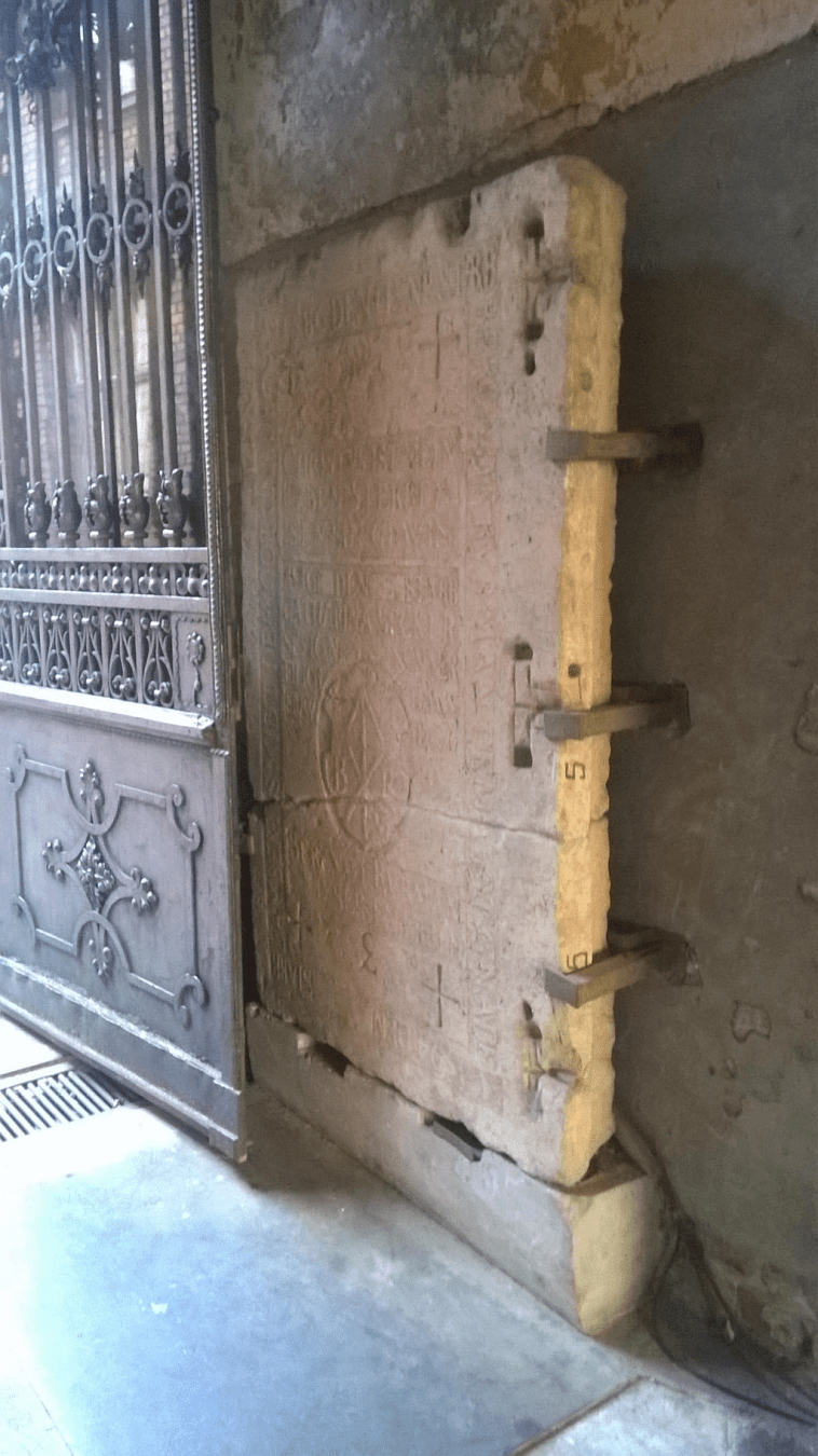 Photo of a stone door, much more faded and broken at the bottom third of the door, with very faded writings across of the stone door. Metal clamps hold the door to the wall.