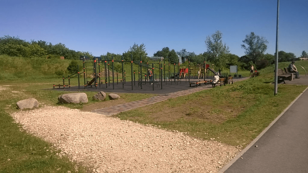 Photo of a new playground in a grassy area by a paved path on a clear summer day, with people hanging around or sitting on the benches.