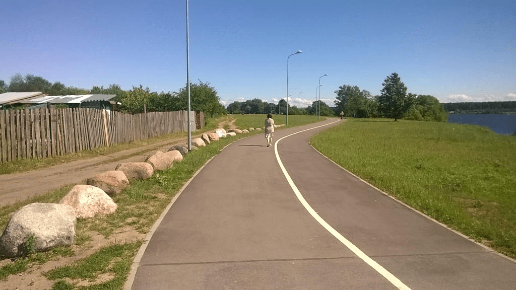 Photo of a paved road through a grassy field, homes on the left side and a grassy field on the right side.