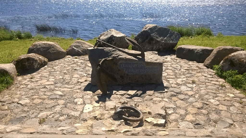 Photo of a rock monument, with a metal anchor embedded in it, with the words "Daugavas anchorers" in Latvian on it, in a rocky spot in a grassy field.