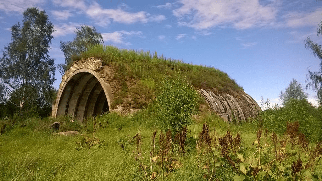 Old hanger, with patchy dirt and grass on it, lush grass around and some stones in said grass