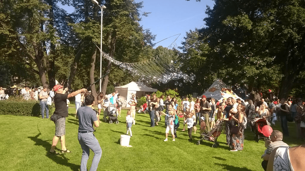 During a summer fair, a man is making bubbles with a makeshift bubble wand of two fishing piles and a long rope soaked in soapy water, a small horde of kids reaching for the bubbles