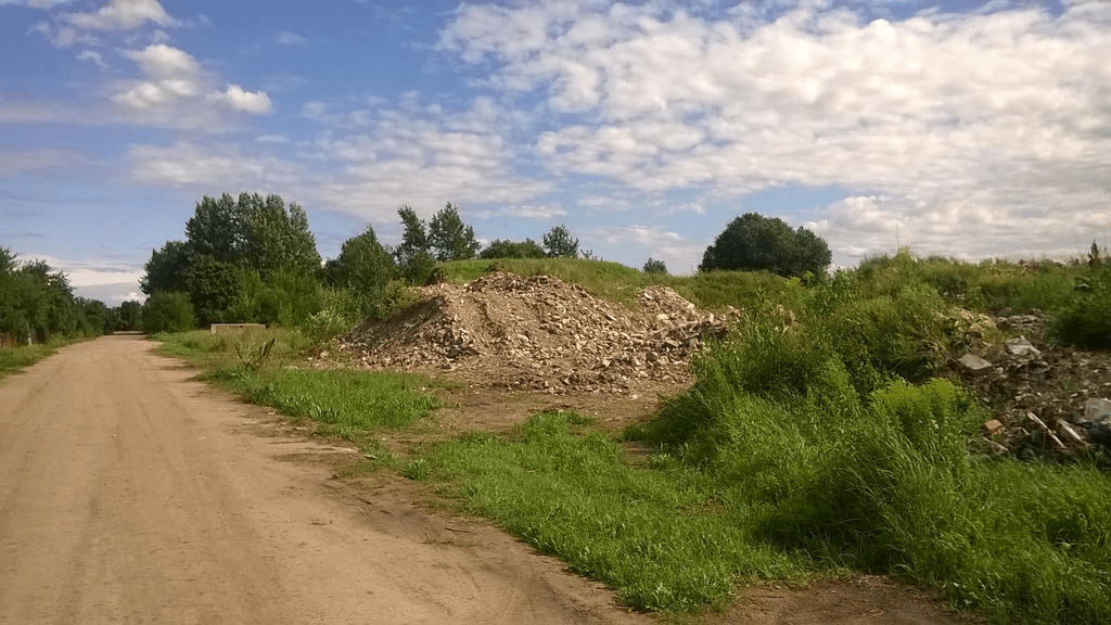 Piles of rocks and stones, with growing grass on them, by a dirt road, leading to a forested area