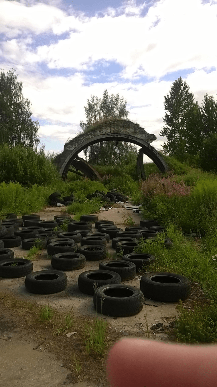 A destroyed old hanger, only remnants are grass covered from arch, with tall grass around and a lot of tires strewn on the road in front of it