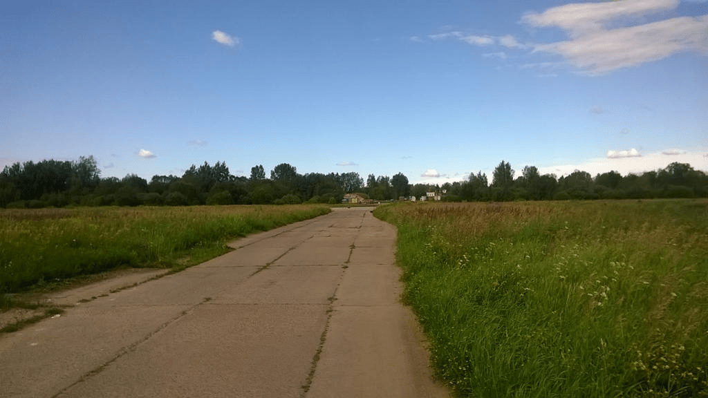 Old taxiway through lush grass, with building in the distance and a line of trees further, the sky bright with some clouds