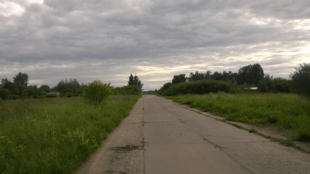 Old road, once a taxiway, through lush grass, bushes and trees, with the sky overcast in blotchy clouds