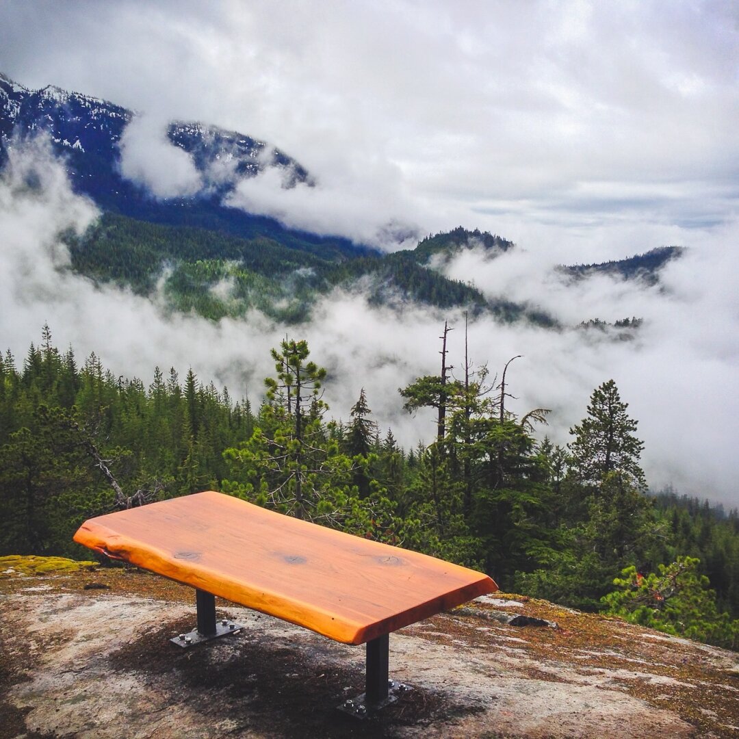 Wooden bench on a rocky outcrop at the Sea to Sky Gondola summit in Squamish, Canada, overlooking misty evergreen forests and snow-dusted mountain peaks under drifting clouds.