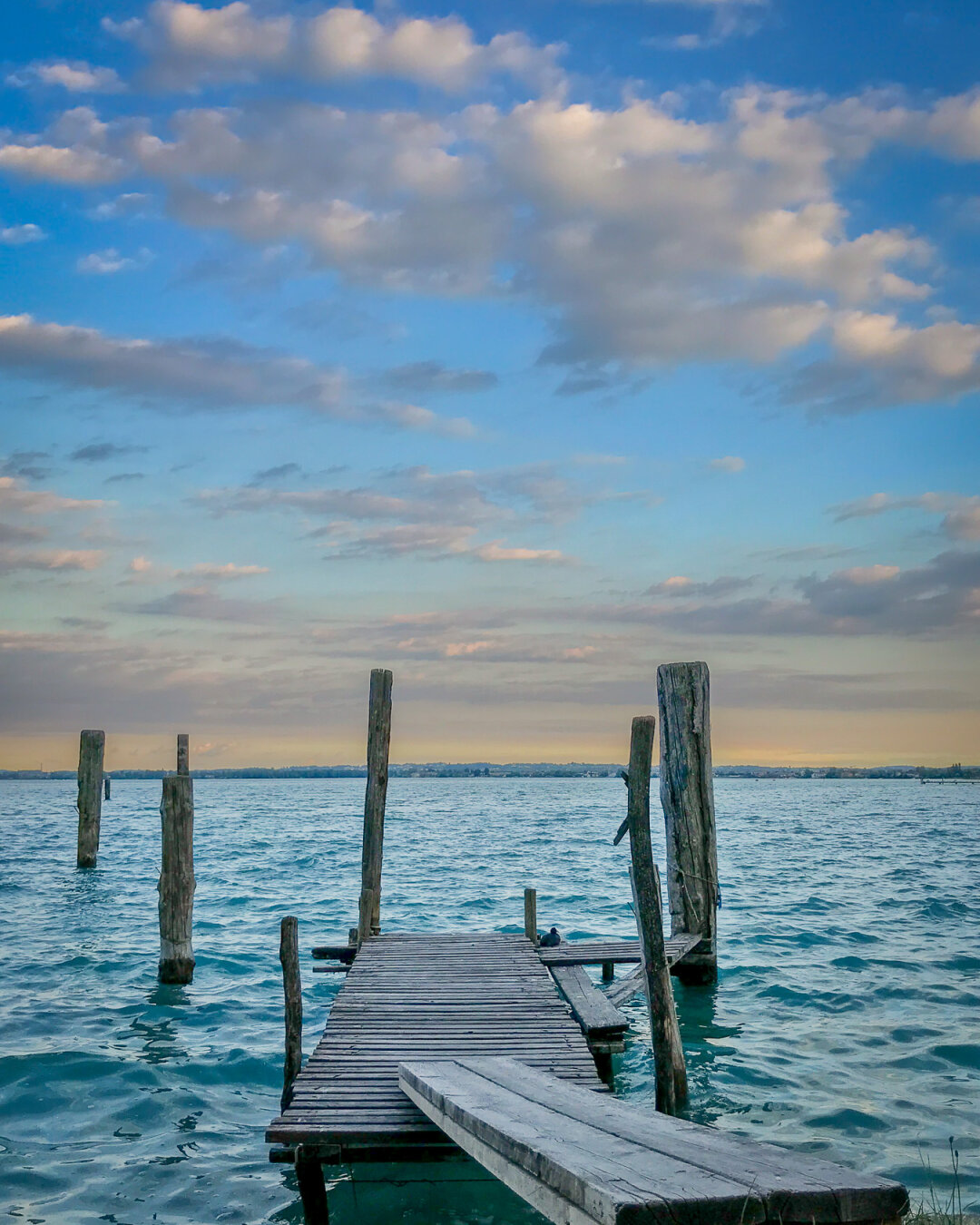 Old wooden pier with missing planks extending into turquoise-blue waters of Lake Garda near Sirmione, Italy, with scattered clouds glowing in the warm light of sunset and distant shoreline in the background