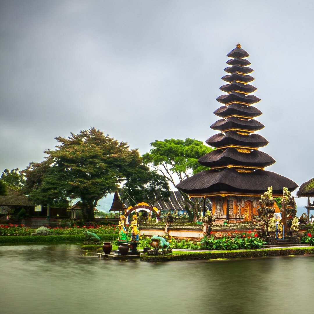 First photo of panorama split in two: Eleven-tiered Meru towers of Pura Ulun Danu Beratan rising from the calm waters of Lake Beratan, shrouded in mist with forested hills and a cloudy sky behind.