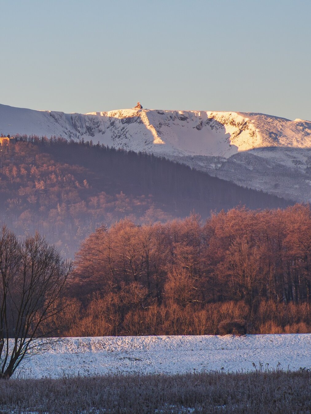 Second half of a split photo:
Chojnik Castle ruins on a snow-covered hill with leafless trees, a snow-filled field in the foreground, and the glacial amphitheaters of Śnieżne Kotły behind under a clear blue sky.