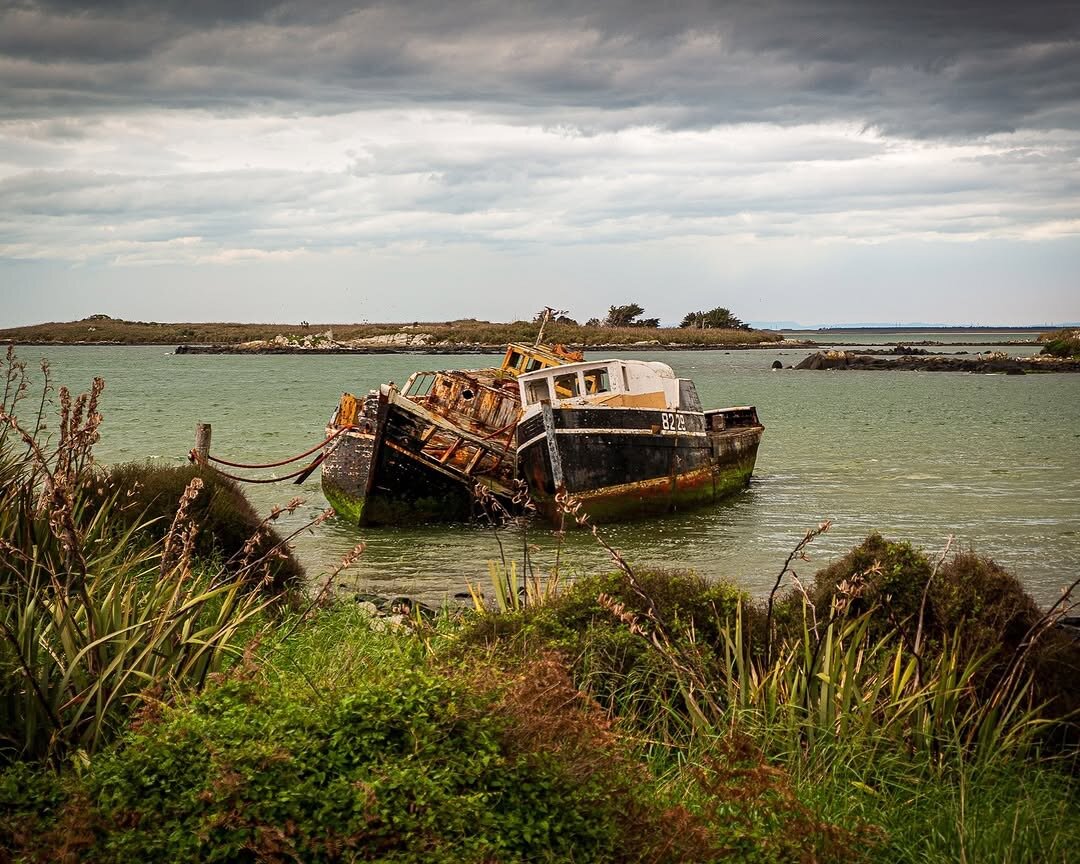 Two rusted, partially submerged boats leaning together at Greenpoint Ship Graveyard near Bluff, South Island, New Zealand, surrounded by seaweed‑lined shallows, green vegetation, and cloudy skies.