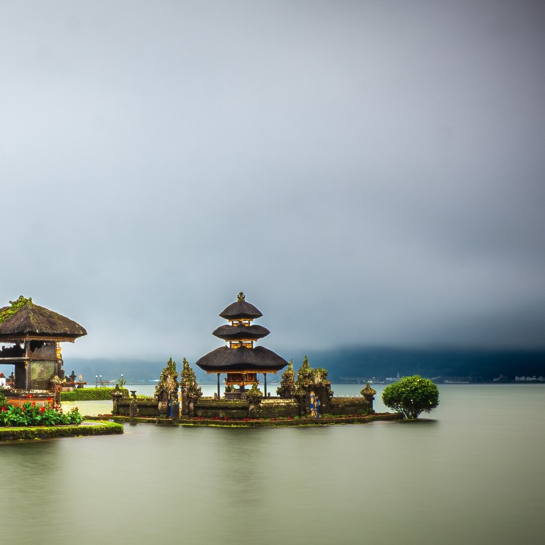 Second photo of panorama split in two: Eleven-tiered Meru towers of Pura Ulun Danu Beratan rising from the calm waters of Lake Beratan, shrouded in mist with forested hills and a cloudy sky behind.