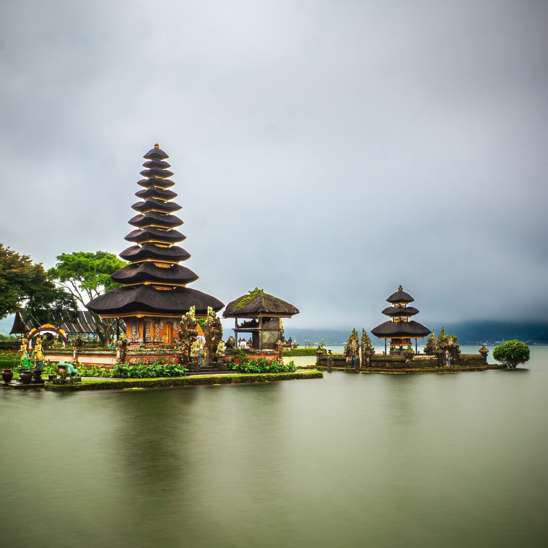 Eleven-tiered Meru towers of Pura Ulun Danu Beratan rising from the calm waters of Lake Beratan, shrouded in mist with forested hills and a cloudy sky behind.